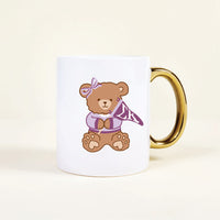 The Sigma Kappa 11 oz Cozy Cute Ceramic Mug by The Darling Effect features a brown teddy bear in a purple bow and sweater, holding an “LK” pennant. The white mug has a gold handle for stylish flair.