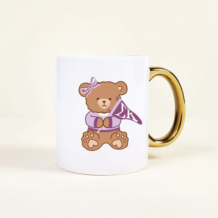 The Sigma Kappa 11 oz Cozy Cute Ceramic Mug by The Darling Effect features a brown teddy bear in a purple bow and sweater, holding an “LK” pennant. The white mug has a gold handle for stylish flair.