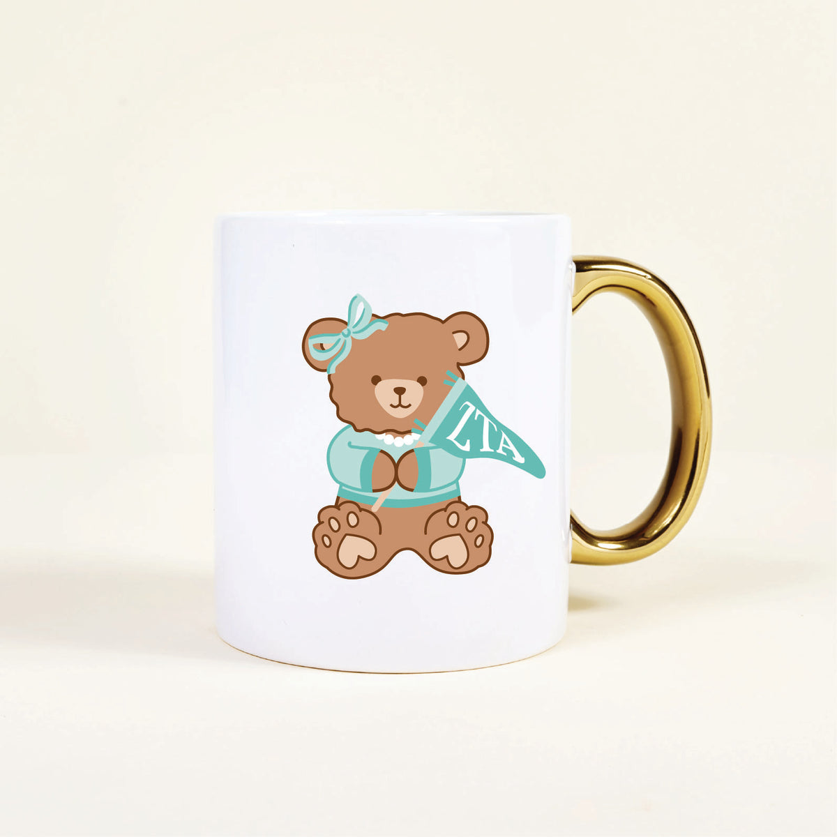 The Darling Effect’s Zeta Tau Alpha 11 oz Cozy Cute Ceramic Mug features a gold handle and an adorable brown teddy bear in a blue bow and sweater holding a teal ZTA pennant—perfect for your morning coffee.