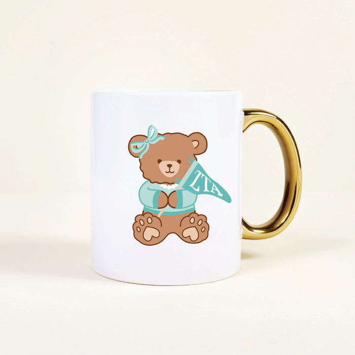 The Darling Effect’s Zeta Tau Alpha 11 oz Cozy Cute Ceramic Mug features a gold handle and an adorable brown teddy bear in a blue bow and sweater holding a teal ZTA pennant—perfect for your morning coffee.