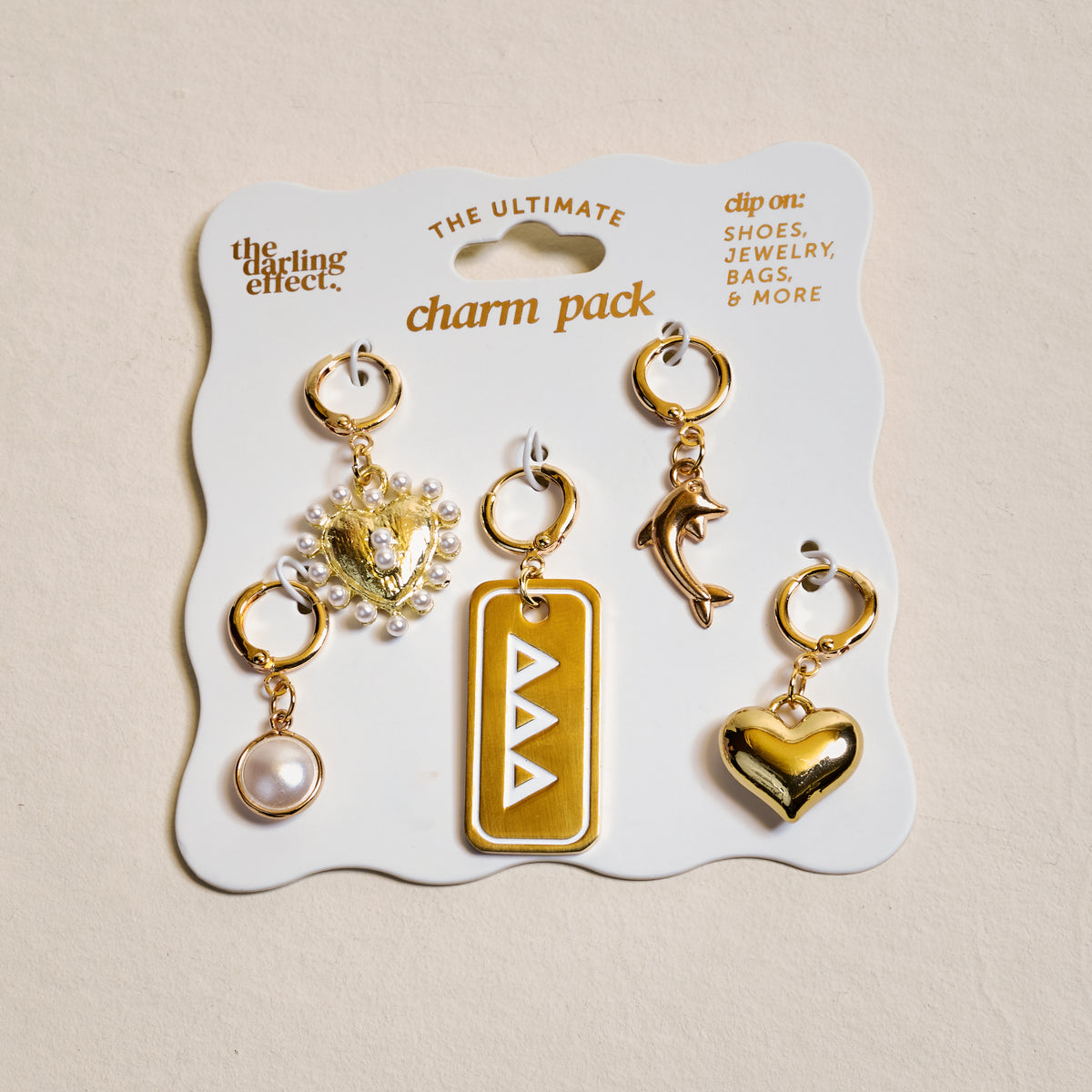 The Darling Effect's Delta Delta Delta The Ultimate Charm Pack features five gold charms—a pearl, jeweled heart, yellow rectangle with white triangles, dolphin, and gold heart—perfect for showing your sorority spirit.