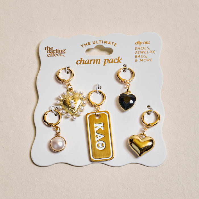 The Darling Effect’s Kappa Alpha Theta The Ultimate Charm Pack features a charm necklace on a white card with five gold charms: a pearl, gold heart with pearls, rectangular "KAΘ" charm, black heart, and shiny gold heart.