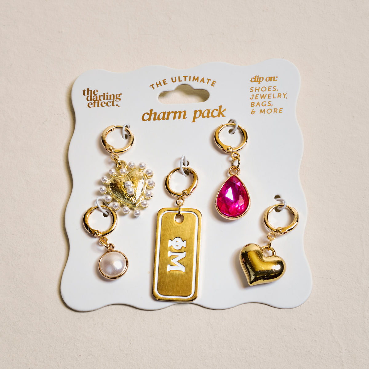 The Darling Effect's Phi Mu The Ultimate Charm Pack features five gold-toned clip-on charms: a beaded crown, “M” tag, pink teardrop gem, pearl, and heart, all presented on a card labeled “the darling effect charm pack.”.
