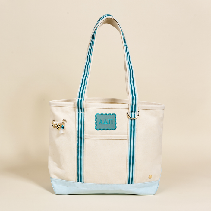 The Darling Effect’s Alpha Delta Pi Canvas Tote Bag features cream canvas, blue striped handles, a light blue base, a gold charm pin on the left, and a blue patch with embroidered sorority letters—perfect for stylish ADPi members.