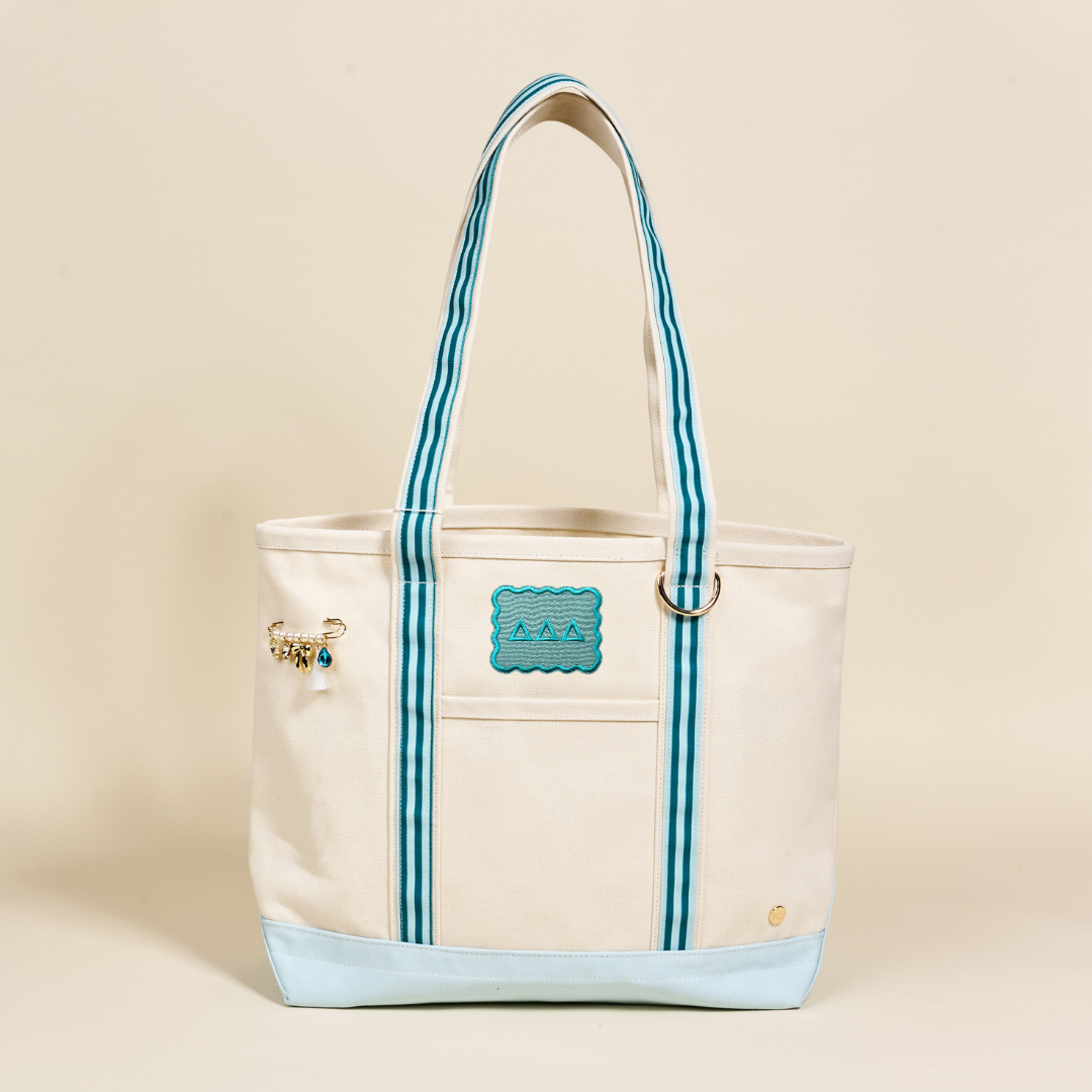 The Delta Delta Delta Canvas Tote Bag by The Darling Effect has cream canvas, blue/white striped handles, a blue wave patch, a gold pin with small charms on the left, and a light blue bottom—perfect for your sorority style.