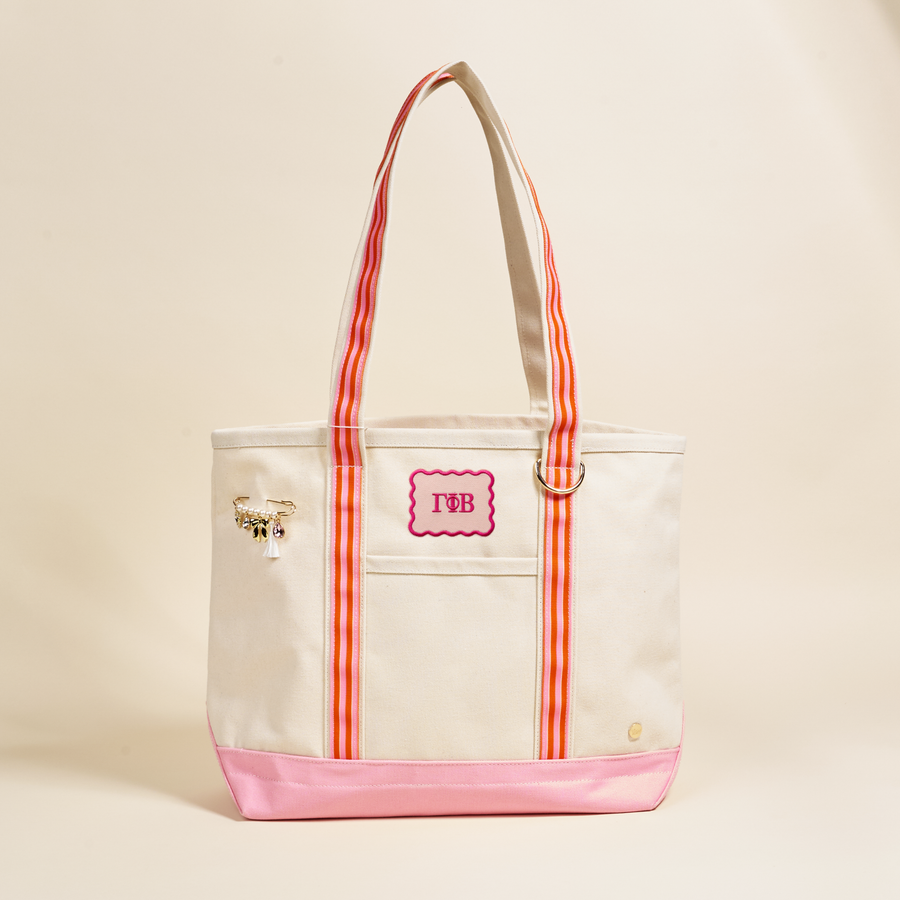 The Darling Effect Gamma Phi Beta Canvas Tote Bag features pink handles, a pink base, red vertical stripes, a pink “TB” monogram patch, and gold charms—ideal as a sorority bag or for everyday style.