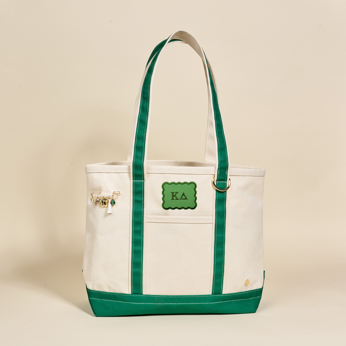 The Darling Effect’s Kappa Delta Canvas Tote Bag features cream canvas, green handles and trim, KA letters on a green patch, a gold charm keychain, and a gold emblem—perfect for everyday sorority use.