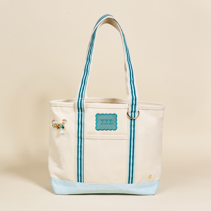 The Sigma Sigma Sigma Canvas Tote Bag by The Darling Effect is a cream-colored, durable canvas tote with light blue accents, striped handles, a blue Greek letters patch, and a decorative charm on the handle. Background: neutral beige.