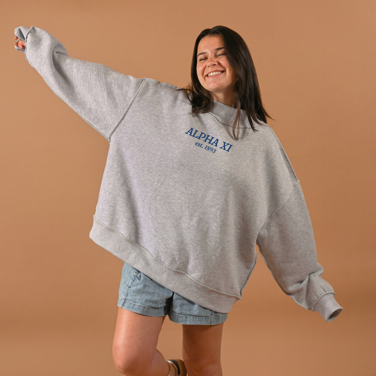 A smiling woman wearing The Darling Effect’s Alpha Xi Delta Chapter Mockneck Sweatshirt S/M and denim shorts stands with arms outstretched against a plain brown background.