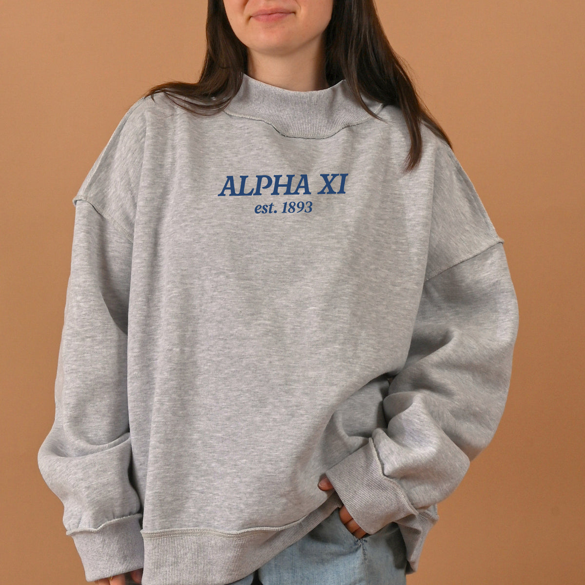 A person is wearing The Darling Effect Alpha Xi Delta Chapter Mockneck Sweatshirt in size S/M, featuring "ALPHA XI est. 1893" in blue text across the chest, standing against a plain tan background.