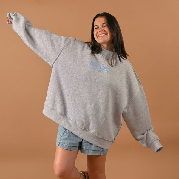 A smiling woman wears The Darling Effect’s Delta Gamma Chapter Mockneck Sweatshirt in S/M with denim shorts, standing against a tan background with arms outstretched in joy.