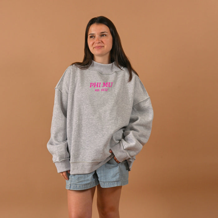 A woman with long dark hair and a slight smile stands against a tan background, wearing The Darling Effect's Phi Mu Chapter Mockneck Sweatshirt L/XL in gray with pink PHI MU est. 1852 lettering, paired with light blue denim shorts.