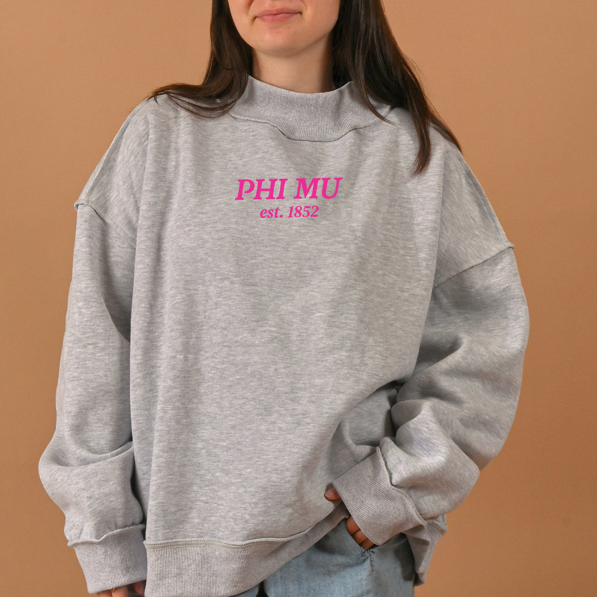 A woman stands against a plain tan background wearing The Darling Effect's Phi Mu Chapter Mockneck Sweatshirt in size S/M—an oversized grey top featuring “PHI MU est. 1852” in pink and blue lettering on the front. This sweatshirt combines comfort and style, making it perfect for any casual occasion.