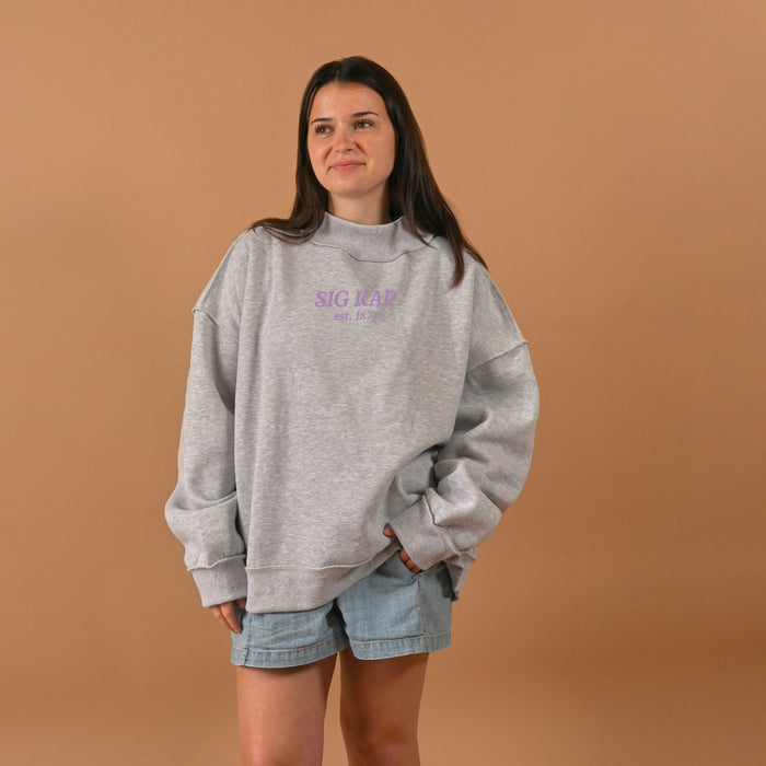 A woman wears The Darling Effect's Sigma Kappa Chapter Mockneck Sweatshirt S/M in gray with purple text and denim shorts, standing before a beige backdrop, smiling slightly with her hands in her pockets.