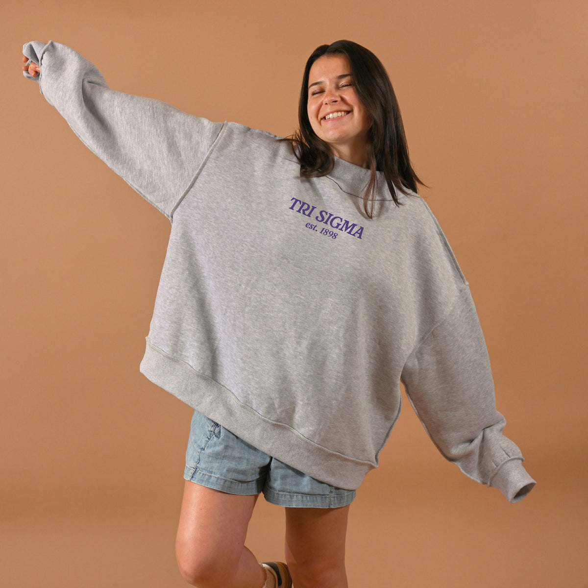 A smiling woman with long dark hair wears The Darling Effect Sigma Sigma Sigma Chapter Mockneck Sweatshirt (S/M) and denim shorts, standing with arms outstretched against a light brown background.