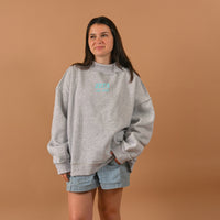 A woman with long dark hair smiles softly, hands in pockets, wearing The Darling Effect's Zeta Tau Alpha Chapter Mockneck Sweatshirt (L/XL) and denim shorts against a plain tan background.