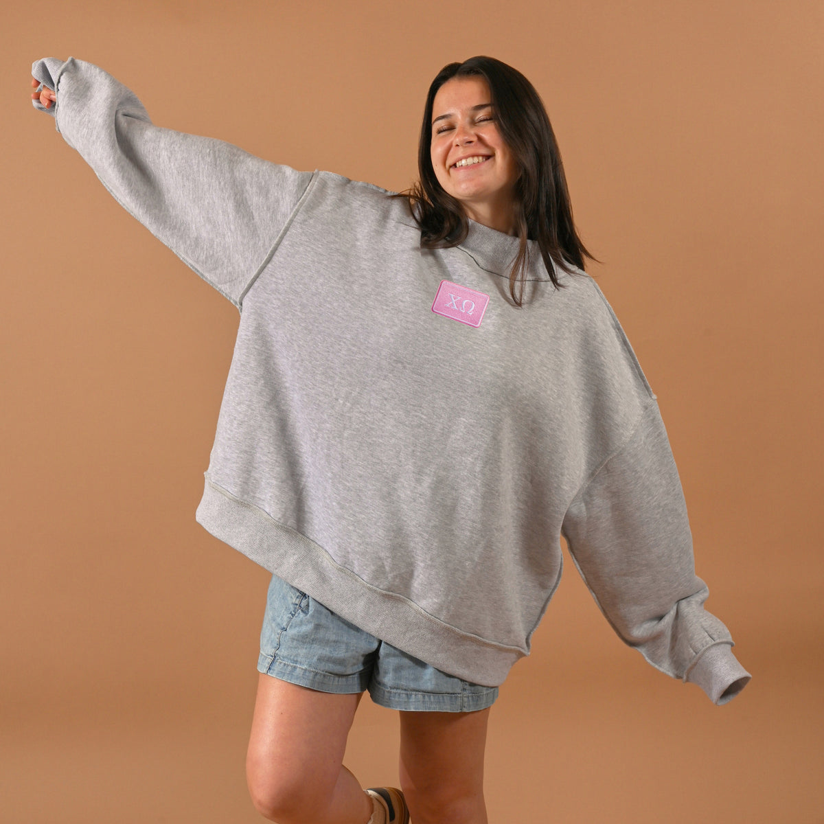 A smiling woman with long dark hair wears The Darling Effect’s oversized gray Chi Omega Letter Patch Mockneck Sweatshirt (S/M) with a pink logo and denim shorts, standing against a tan background with arms outstretched.