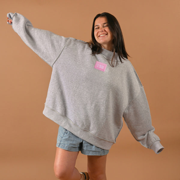 A smiling woman in a gray Gamma Phi Beta Letter Patch Mockneck Sweatshirt by The Darling Effect (L/XL) and denim shorts stands with arms outstretched against a plain brown background.