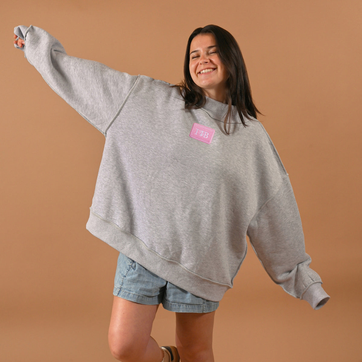 A woman smiles with her eyes closed and arms outstretched, wearing The Darling Effect's Gamma Phi Beta Letter Patch Mockneck Sweatshirt in S/M, paired with denim shorts against a plain beige background.