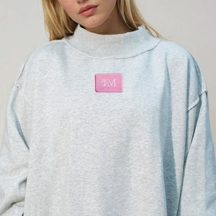 A person with blonde hair wears The Darling Effect Phi Mu Letter Patch Mockneck Sweatshirt in light gray, L/XL, featuring a pink patch with white ΦM letters on the chest. The mock neck frames their partially visible face.