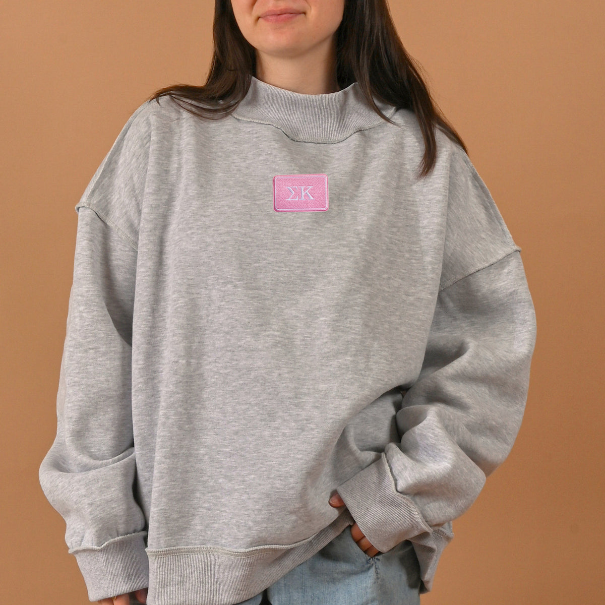 A woman wearing The Darling Effect’s Sigma Kappa Letter Patch Mockneck Sweatshirt in gray (size S/M), featuring a pink Greek letters patch, stands against a plain brown background.