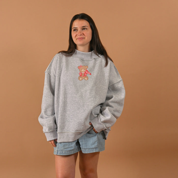 A woman with long brown hair and a subtle smile stands against a beige background, wearing The Darling Effect's Alpha Gamma Delta Teddy Bear Mockneck Sweatshirt in L/XL and light denim shorts.