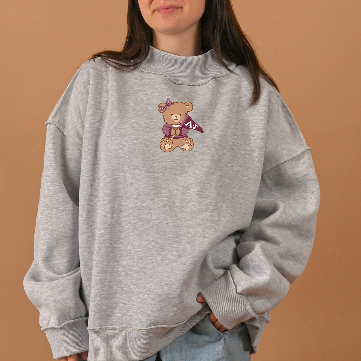 A person wearing The Darling Effect's Alpha Phi Teddy Bear Mockneck Sweatshirt in gray, size L/XL—featuring a cute bear graphic holding a purple flag and a gift box—stands in front of a plain tan background.