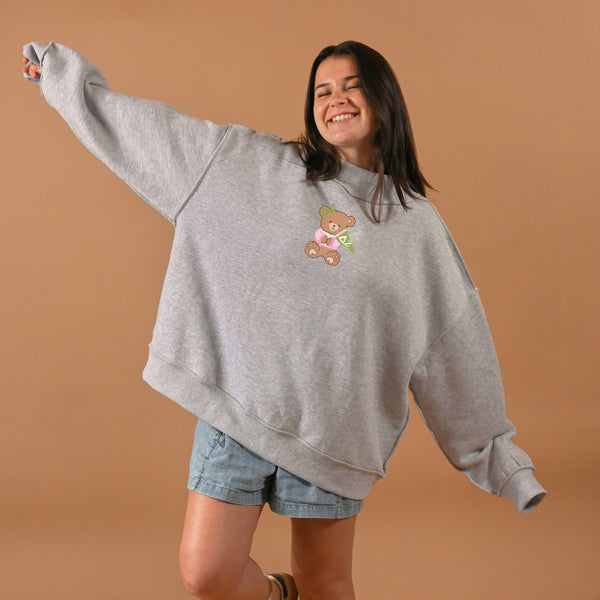 A smiling woman poses playfully in The Darling Effect's Delta Zeta Teddy Bear Mockneck Sweatshirt (S/M) and denim shorts, standing against a plain beige background.