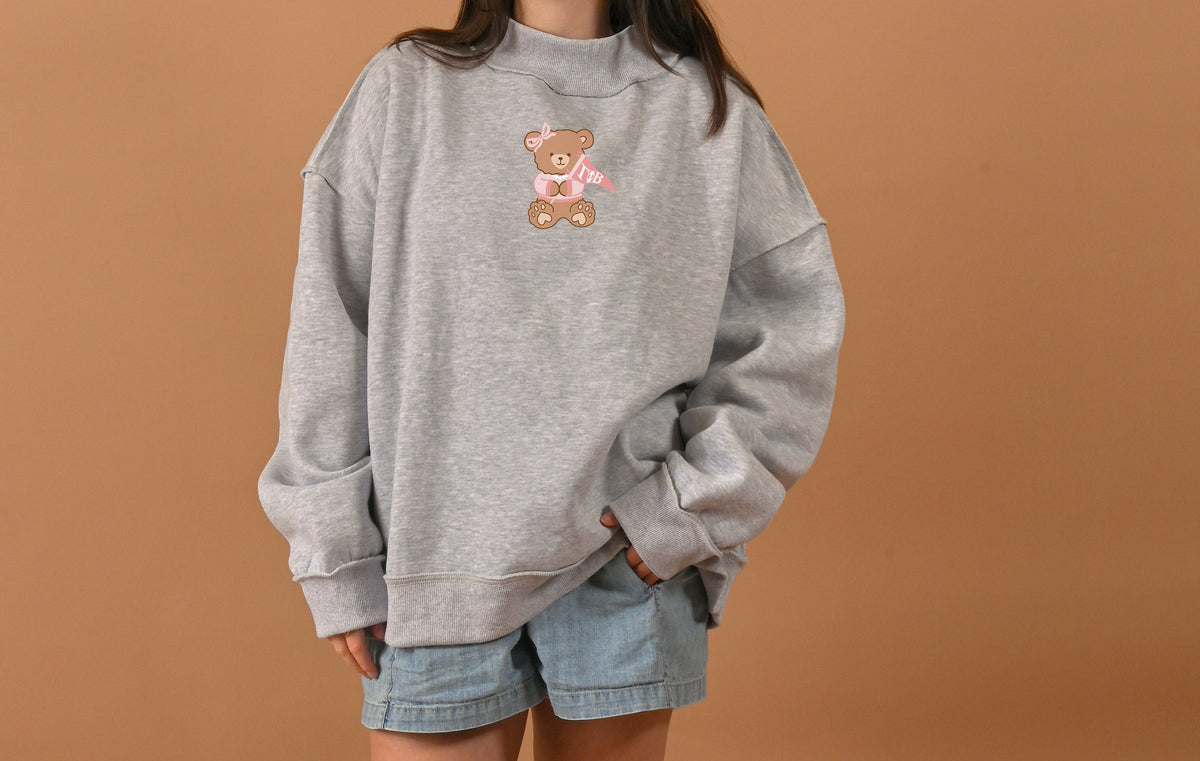 Someone wearing The Darling Effect's Gamma Phi Beta Teddy Bear Mockneck Sweatshirt (S/M) in gray and light blue denim shorts stands against a plain beige background.