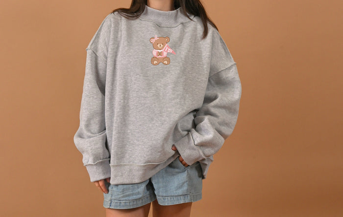 Someone wearing The Darling Effect's Gamma Phi Beta Teddy Bear Mockneck Sweatshirt (S/M) in gray and light blue denim shorts stands against a plain beige background.