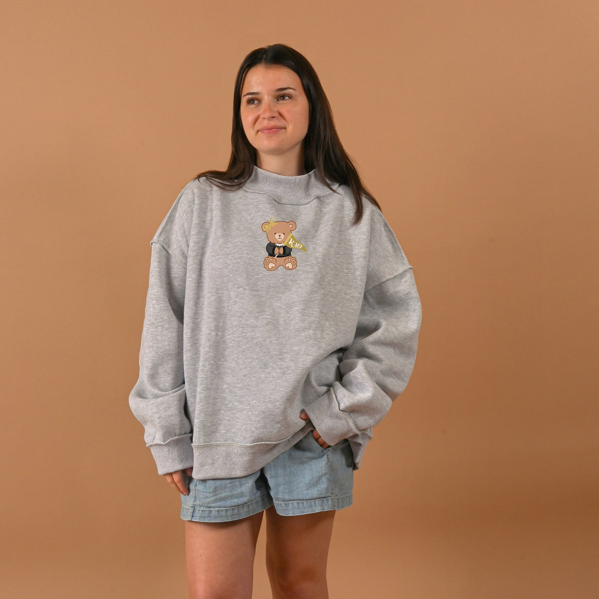 A woman with long brown hair and a slight smile stands against a tan background, wearing The Darling Effect Kappa Alpha Theta Teddy Bear Mockneck Sweatshirt (L/XL) and light blue denim shorts.