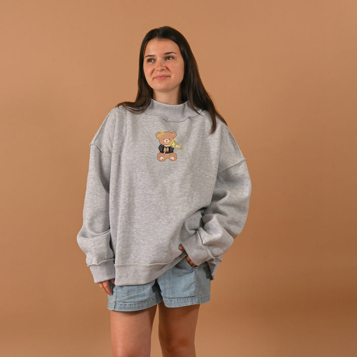 A woman with long brown hair stands against a beige background, wearing The Darling Effect's Kappa Alpha Theta Teddy Bear Mockneck Sweatshirt (S/M) in gray and light denim shorts, hands in pockets and a relaxed expression.