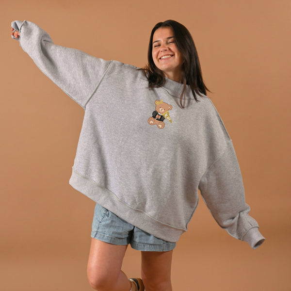A smiling woman poses with arms outstretched in The Darling Effect’s Kappa Alpha Theta Teddy Bear Mockneck Sweatshirt (S/M) and denim shorts against a plain brown background.