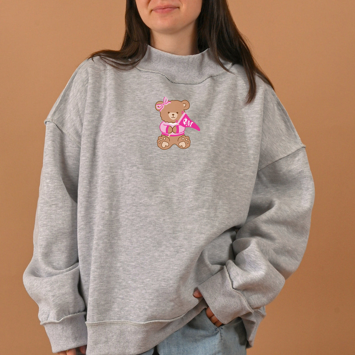 A person wearing The Darling Effect's Phi Mu Teddy Bear Mockneck Sweatshirt in size L/XL stands against a plain beige background; the gray sweatshirt features a cute teddy bear graphic holding a pink heart and wearing a bow.