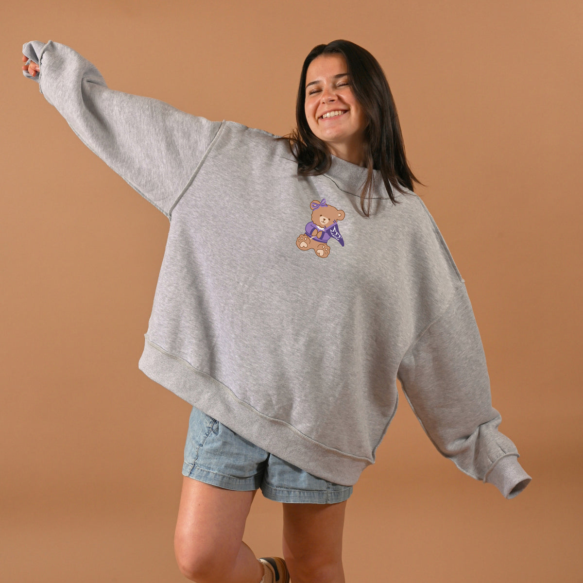 A smiling woman poses with arms outstretched in front of a light brown background, wearing The Darling Effect’s Sigma Sigma Sigma Teddy Bear Mockneck Sweatshirt (size S/M) and denim shorts.