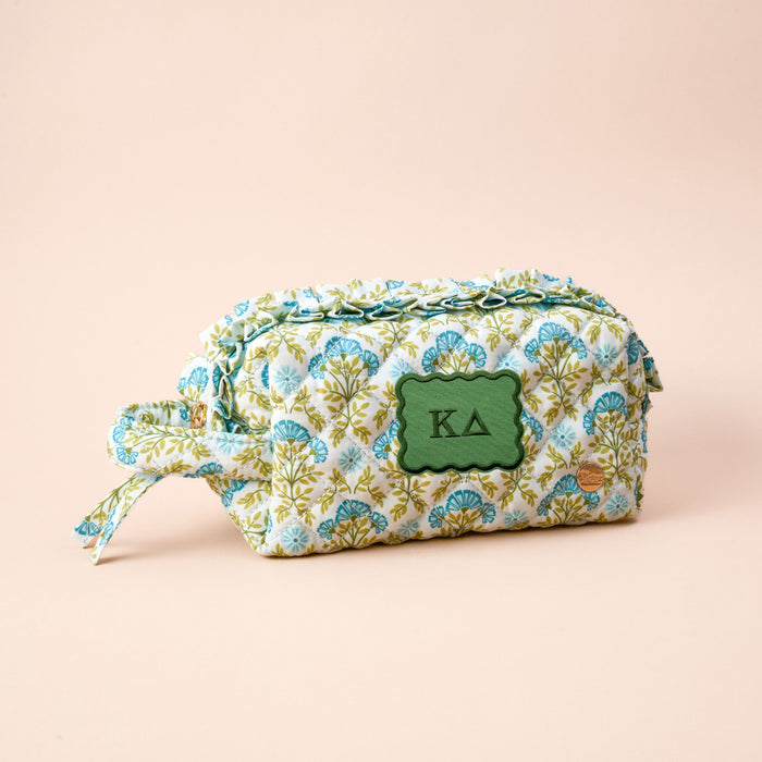 The Darling Effect's Kappa Delta Essentials Quilted Cosmetic Pouch features a blue and green floral design, a green KA patch on the front, and a side strap—ideal for your sorority essentials. Shown on a light beige background.