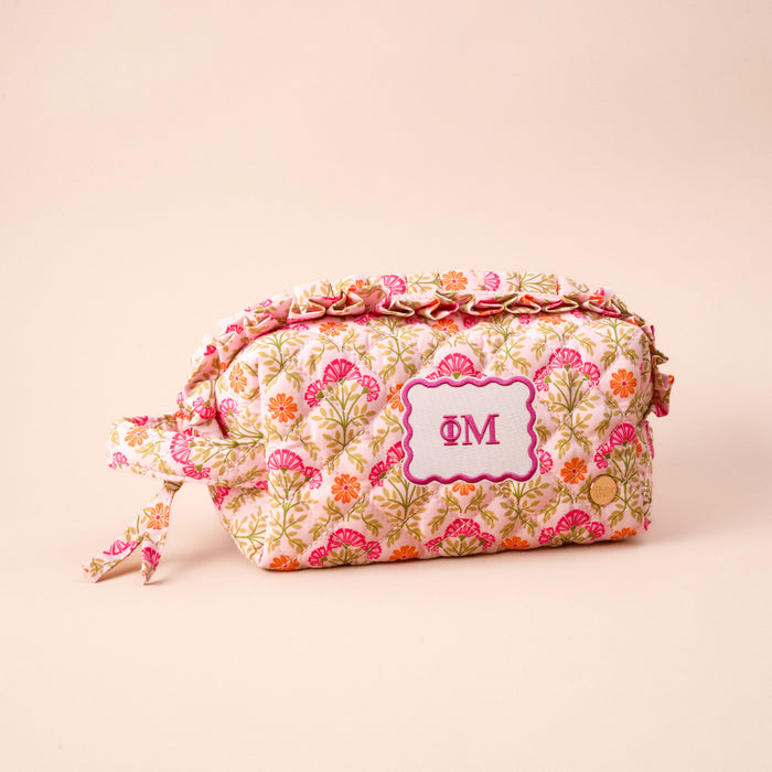 The Darling Effect's Phi Mu Essentials Quilted Cosmetic Pouch features a floral pattern, side tie, and stitched white label with pink "ΦΜ" Greek letters on a soft light beige background.