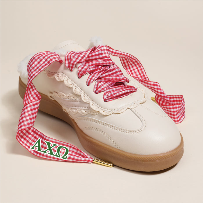 Alpha Chi Omega Step It Up Shoelaces from The Darling Effect feature red gingham laces with green and red ΑΧΩ Greek letters on the aglet—ideal for unique sorority gifts. Shown on a white sneaker with a gum sole and lace trim against a beige background.
