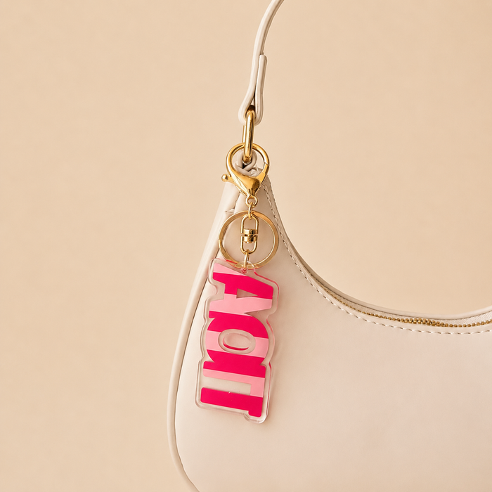 A close-up of a white handbag with a gold clasp and The Darling Effect’s Alpha Omicron Pi Stripes Acrylic Keychain attached. The pink and white layered letters spell out "LOVE." The soft beige background adds a gentle touch. Perfect sorority gift!.