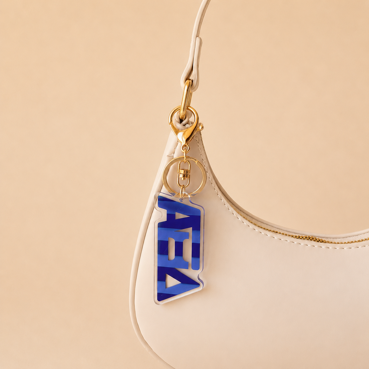 A beige handbag with gold hardware displays The Darling Effect’s Alpha Xi Delta Stripes Acrylic Keychain in blue and clear on the strap, featuring stylized Greek letters. The beige background makes it a perfect stylish sorority gift.