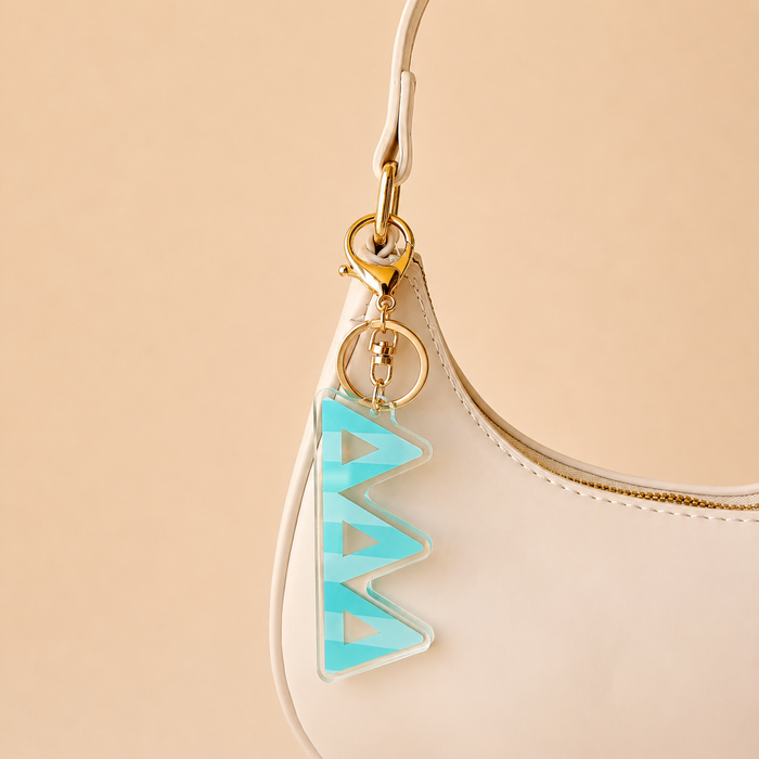 A cream-colored handbag with a gold clasp features The Darling Effect’s Delta Delta Delta Stripes Acrylic Keychain in turquoise and clear zigzags on the handle—perfect as a stylish bid day gift. The background is plain light beige.