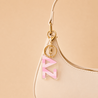 A cream-colored handbag with a gold keychain on the strap, featuring The Darling Effect’s Delta Zeta Stripes Acrylic Keychain—perfect as a chic sorority gift or bid day accessory against a beige background.