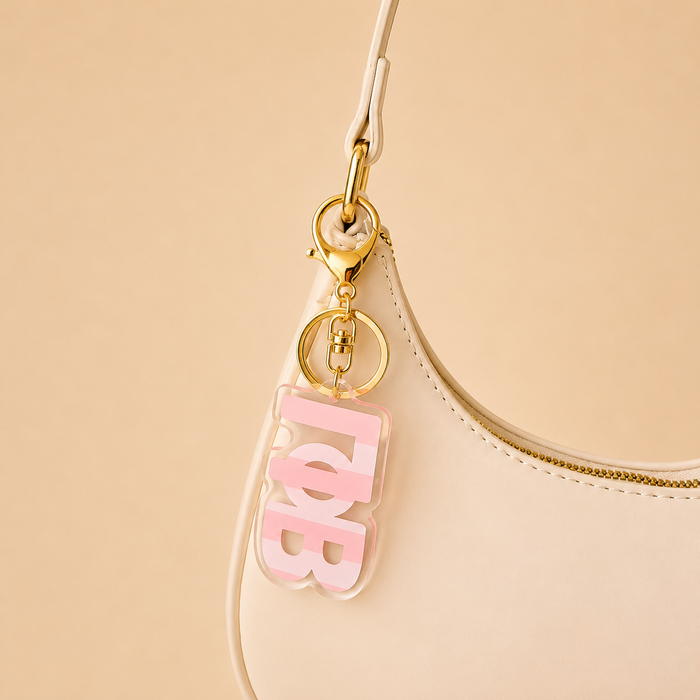 A cream-colored handbag with a gold zipper features The Darling Effect's Gamma Phi Beta Stripes Acrylic Keychain in pink and white—an ideal sorority gift.