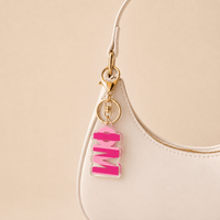 A close-up of a white handbag with a gold clasp holding The Darling Effect’s Phi Mu Stripes Acrylic Keychain—shaped like the word MOM in block letters. The background is a soft beige, making it a perfect sorority gift.