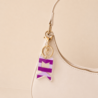 A close-up of a white handbag with a gold zipper and The Darling Effect's Sigma Kappa Stripes Acrylic Keychain, featuring bold purple and white geometric stripes—an eye-catching sorority gift.