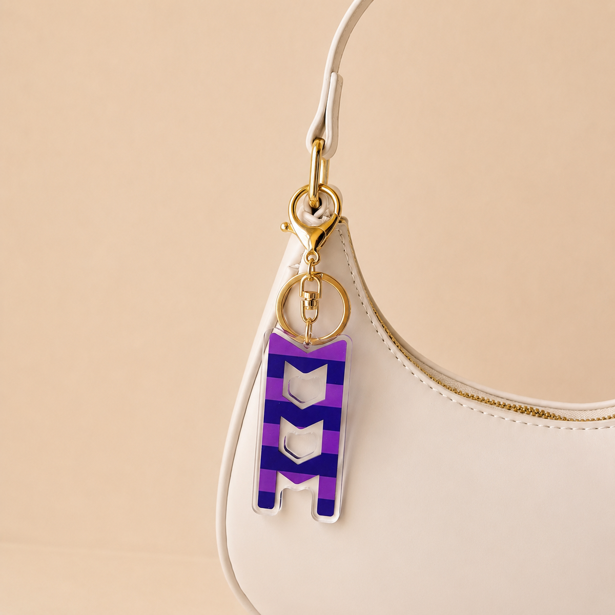 A white handbag with gold hardware showcases The Darling Effect’s Sigma Sigma Sigma Stripes Acrylic Keychain, shaped like an M in purple and white stripes and attached to the strap, against a soft beige background—an ideal sorority gift.