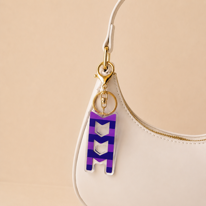 A white handbag with gold hardware showcases The Darling Effect’s Sigma Sigma Sigma Stripes Acrylic Keychain, shaped like an M in purple and white stripes and attached to the strap, against a soft beige background—an ideal sorority gift.