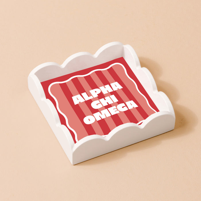 The Darling Effect’s Alpha Chi Omega Scallop Trinket Tray is a square, white tray with scalloped edges and a red and pink striped insert that reads “ALPHA CHI OMEGA”—perfect for holding jewelry or keepsakes.