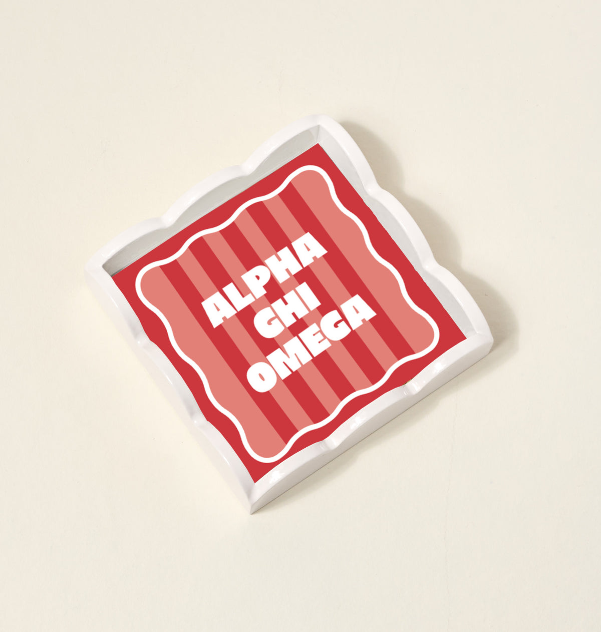 The Darling Effect Alpha Chi Omega Scallop Trinket Tray features a square, wavy-edged design with red and pink stripes and bold white text, perfect as an accessory or jewelry organizer.