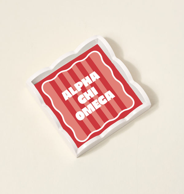 The Darling Effect Alpha Chi Omega Scallop Trinket Tray features a square, wavy-edged design with red and pink stripes and bold white text, perfect as an accessory or jewelry organizer.